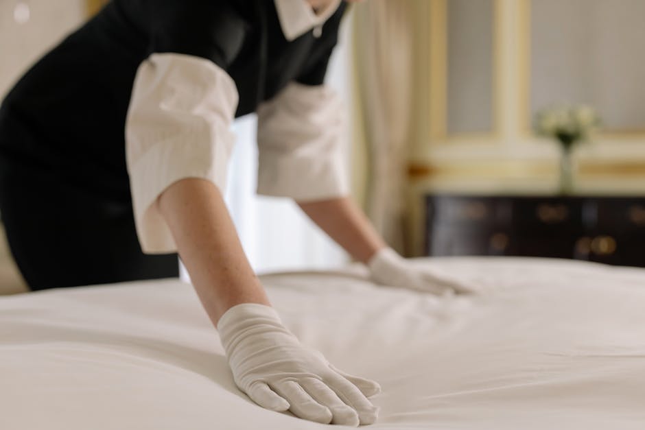 A person wearing white gloves and a white shirt is carefully placing a decorative beige pillow with a subtle geometric pattern onto a white sofa. The sofa has a tufted beige headboard and is decorated