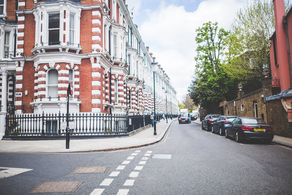 A quiet residential street featuring a row of Victorian-style terraced houses with red brick and white decorative trim, set behind black wrought iron fencing. The street has parked cars along the side