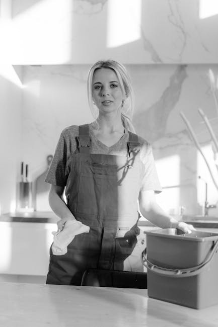 A young woman with light-colored hair, dressed in a casual t-shirt and overalls, stands in a modern kitchen holding a cleaning cloth in her right hand and resting her left hand on a grey plastic bucke