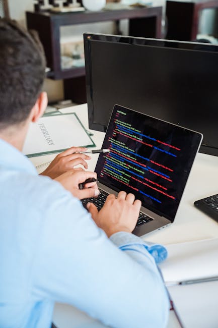 A close-up photograph of a laptop displaying lines of code on its screen, situated on a wooden desk. Next to the laptop, there is a ceramic mug with a beige background and black text that reads 'THIS