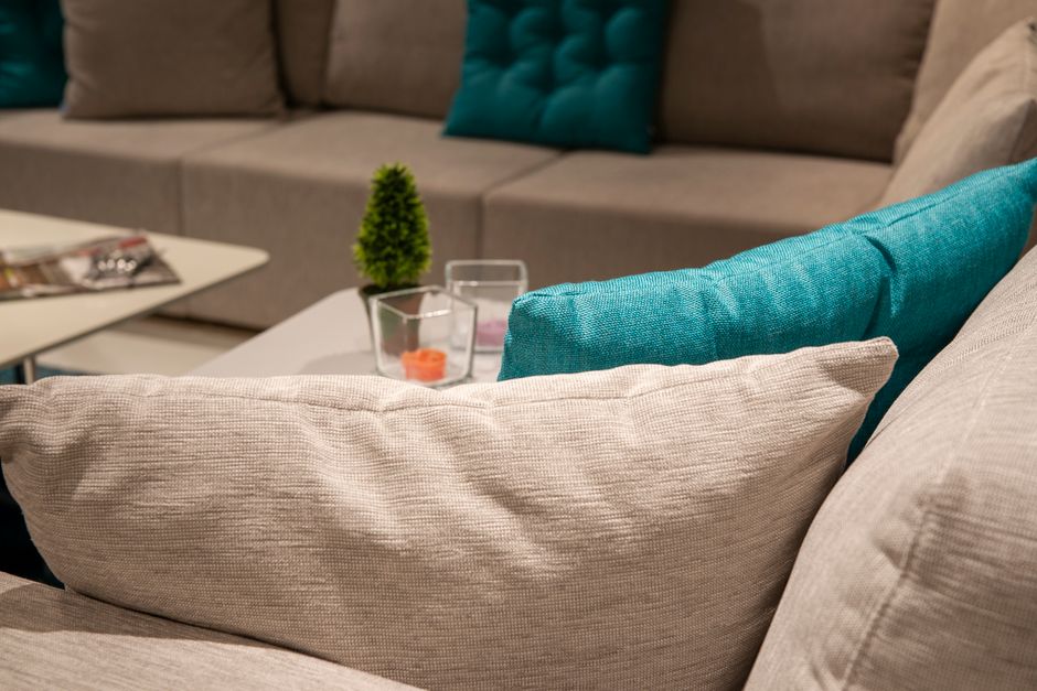 Close-up view of a beige fabric sofa in a living room, featuring neatly arranged teal and beige cushions. The surface of the sofa appears clean and well-maintained, with no visible dust or stains. In
